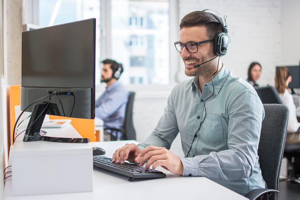Smiling handsome male customer support phone operator with headset working in call centre. Group of sales agent working in office.