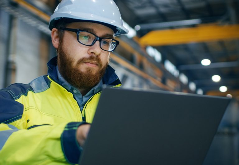 Industrial Engineer in Hard Hat Wearing Safety Jacket Uses Touchscreen Laptop. He Works at the Heavy Industry Manufacturing Factory. In the Background Welding/ Metalworking Processes are in Progress.